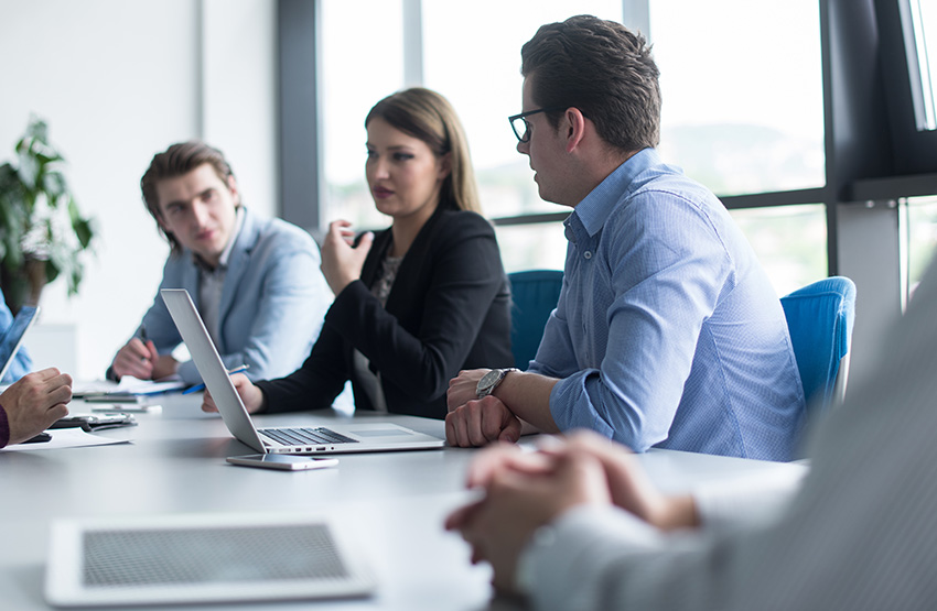 A group of professionals engaged in a discussion around a large table in a bright, modern office. The setting includes laptops, a tablet, and natural light streaming through large windows, emphasizing collaboration and teamwork.